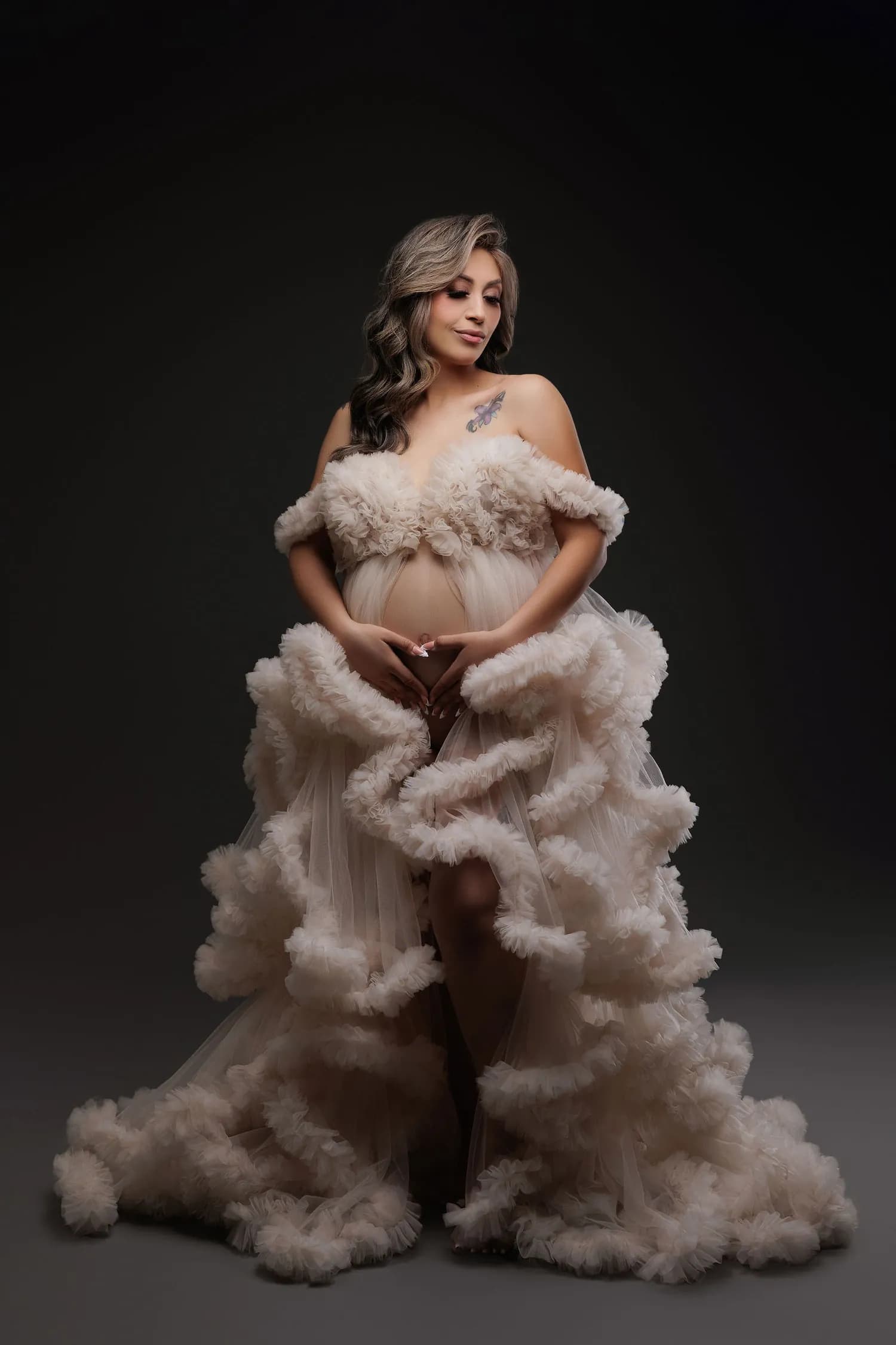Pregnant woman in a blush ruffled tulle gown against a dark backdrop
