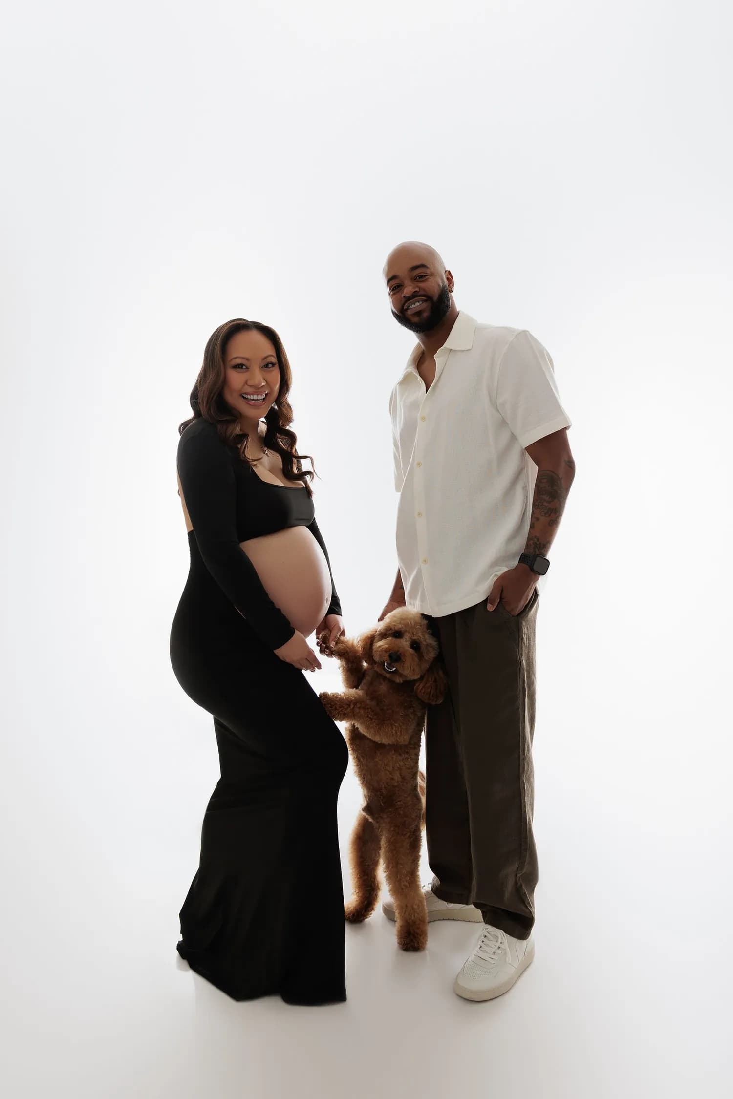 Expecting couple on a white background with a small curly-haired dog standing up against her