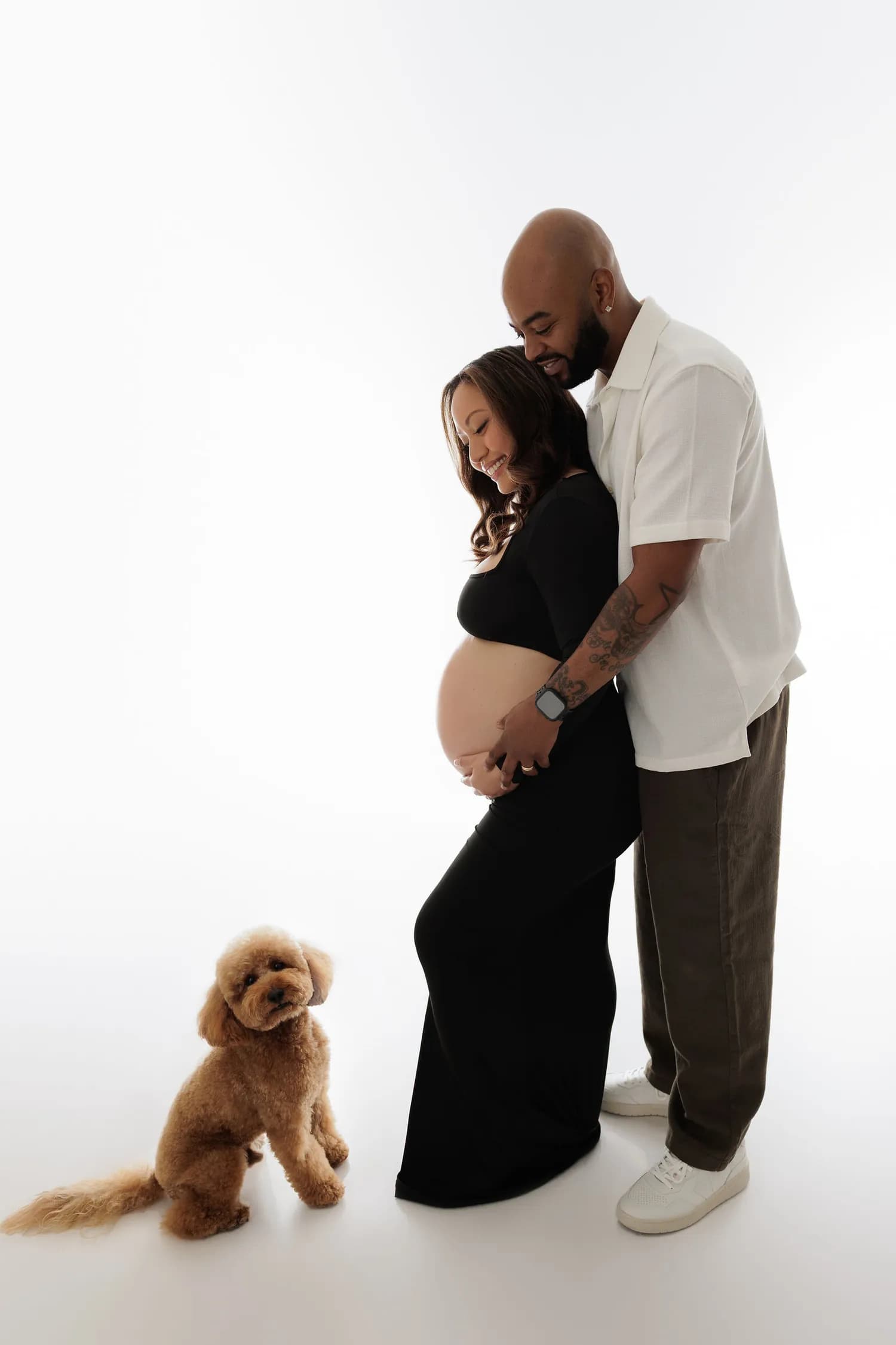 Expecting couple embracing while a small curly-haired dog sits beside them