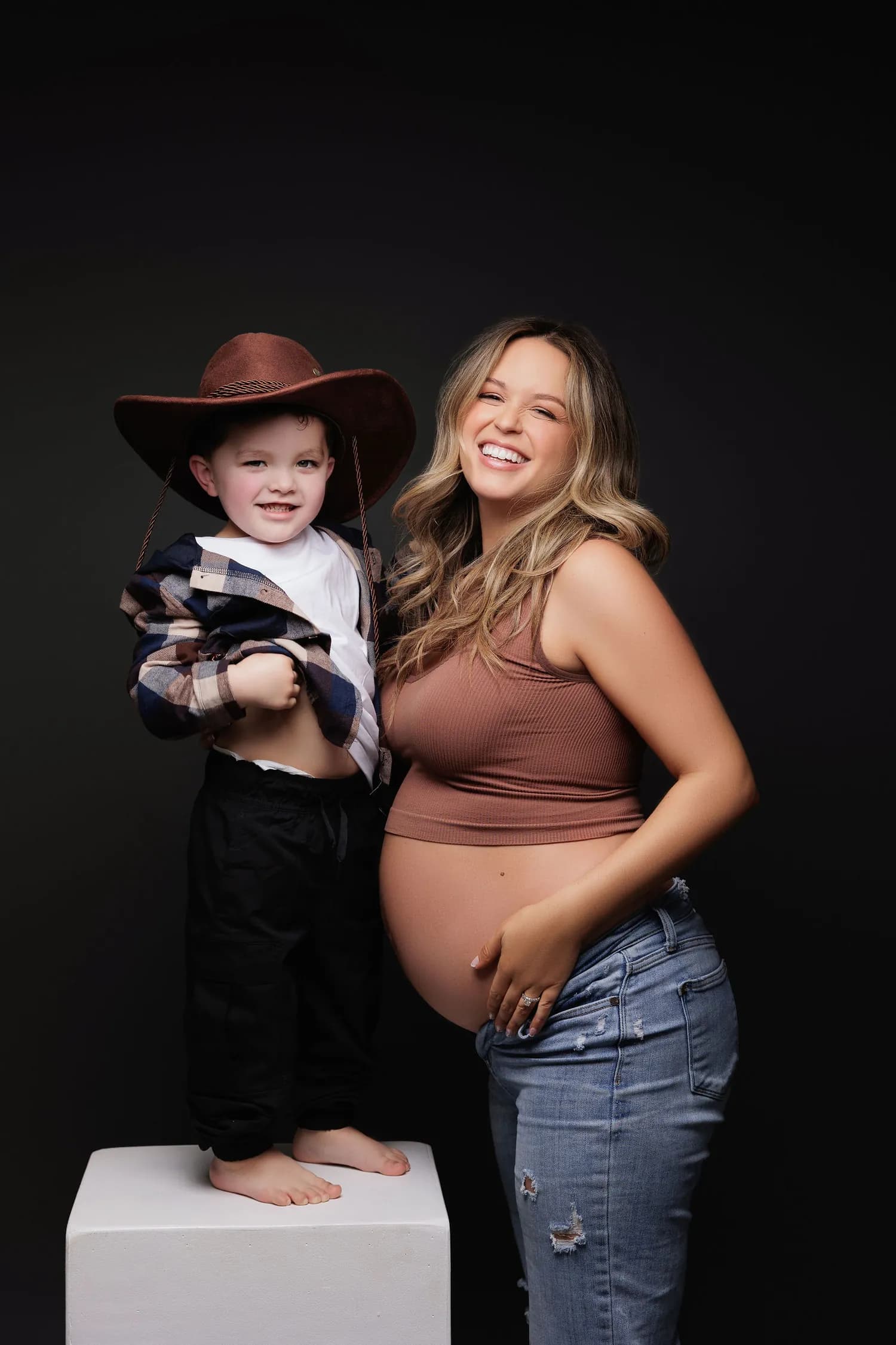 Laughing pregnant mother beside a little boy in a cowboy hat standing on a white box
