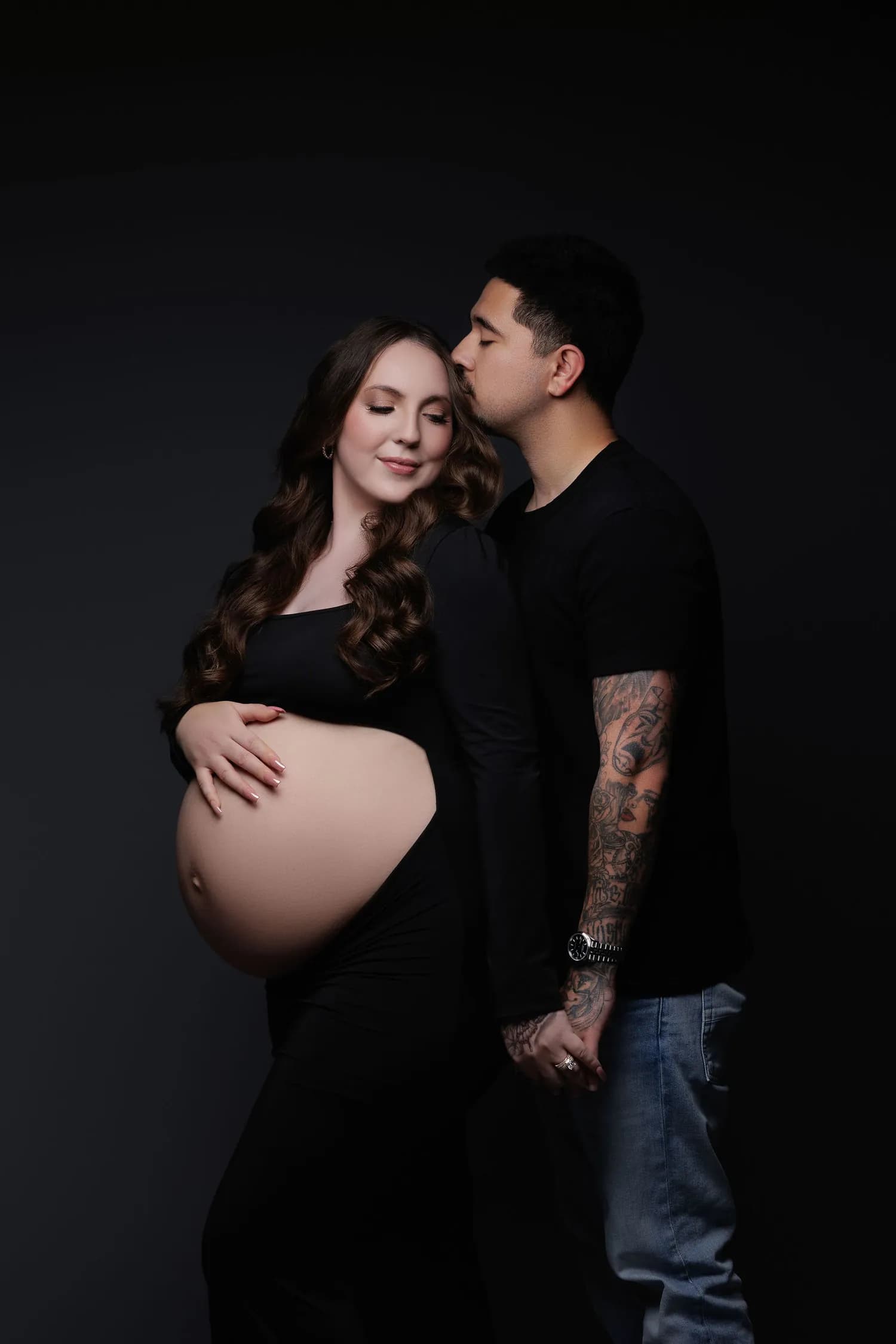 Partner kissing the forehead of a pregnant woman in black against a dark backdrop