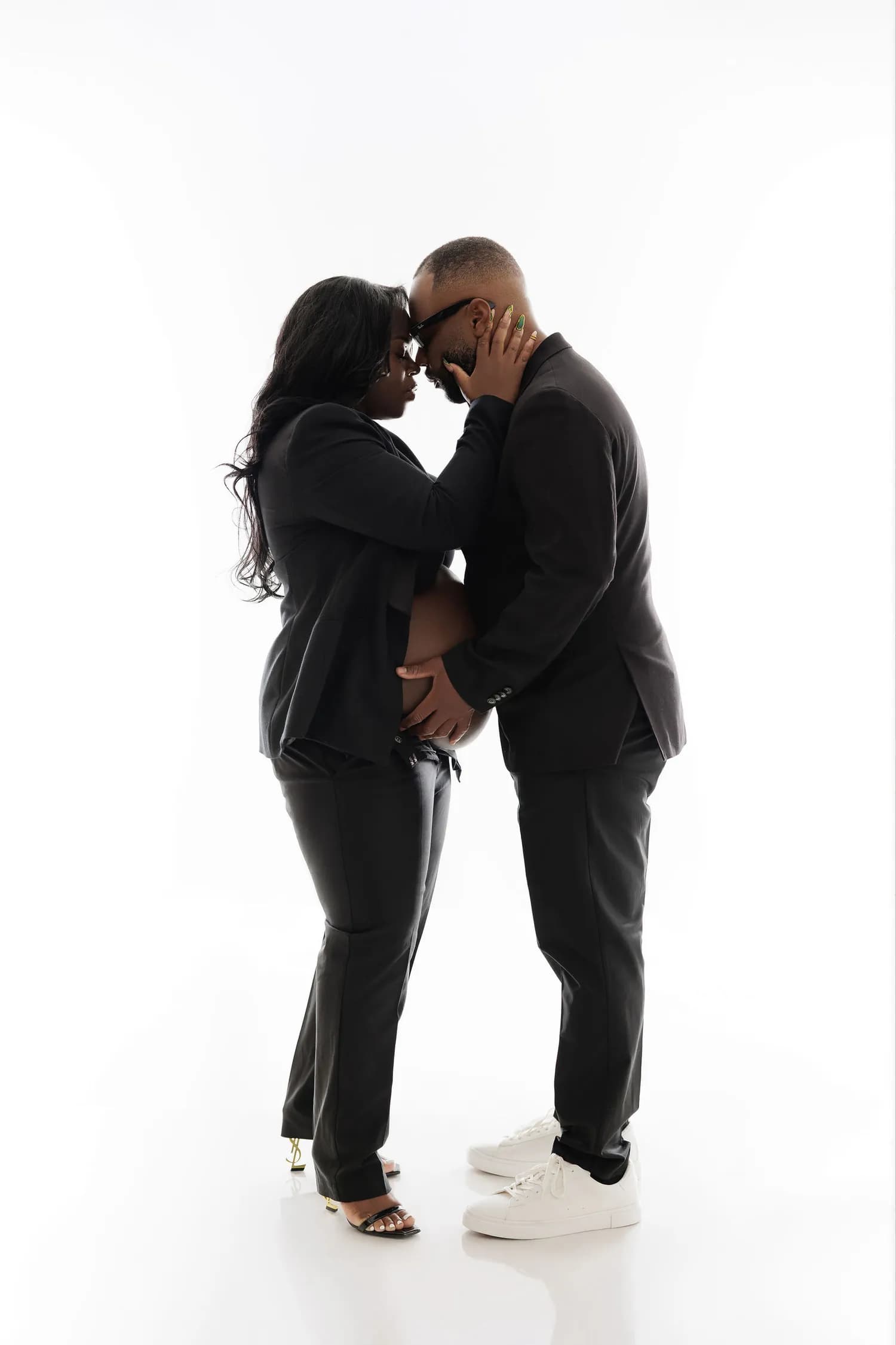 Expecting couple in black suits with their foreheads together on a white background