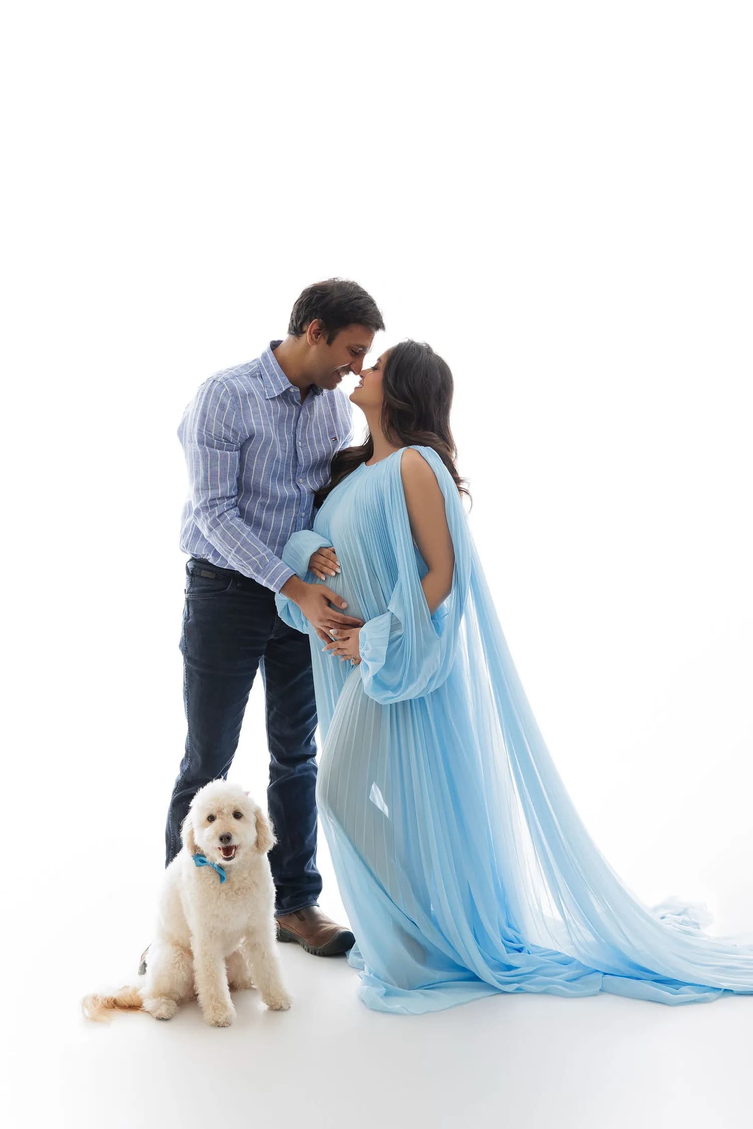 Couple nose to nose, the pregnant woman in pale blue chiffon, with a white dog at their feet