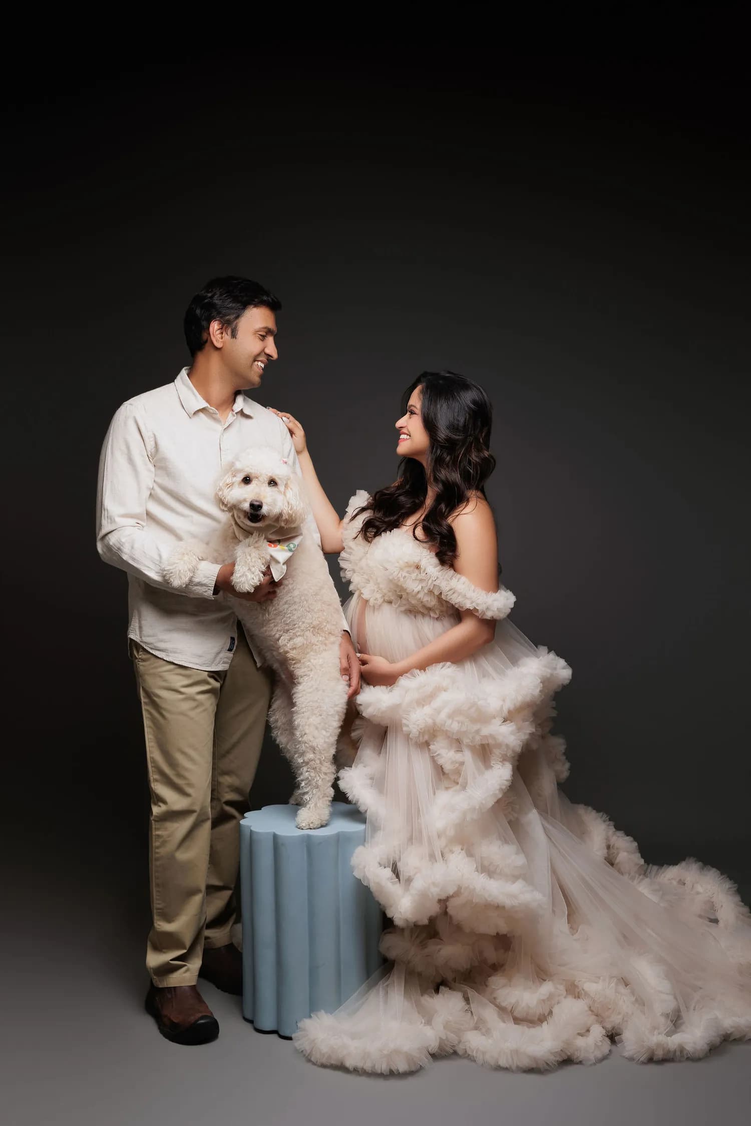 Couple holding a white dog, the pregnant woman in a blush ruffled gown, against a grey backdrop