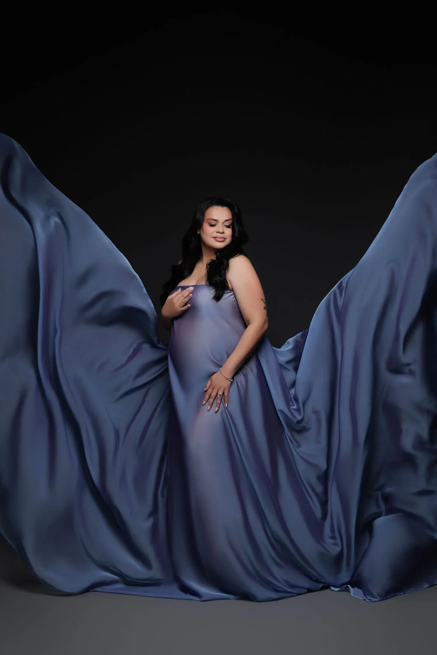 Pregnant woman in blue satin with the fabric spread wide around her against a dark backdrop