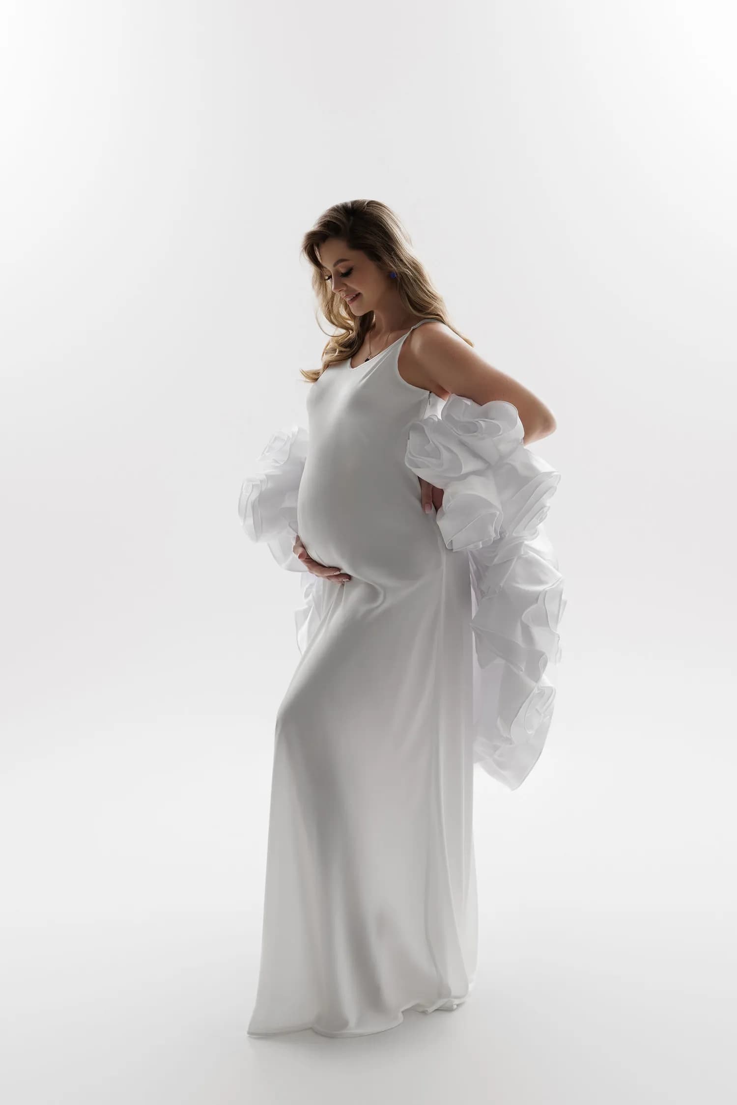 Pregnant woman in a white satin slip dress with ruffled sleeves on a white background