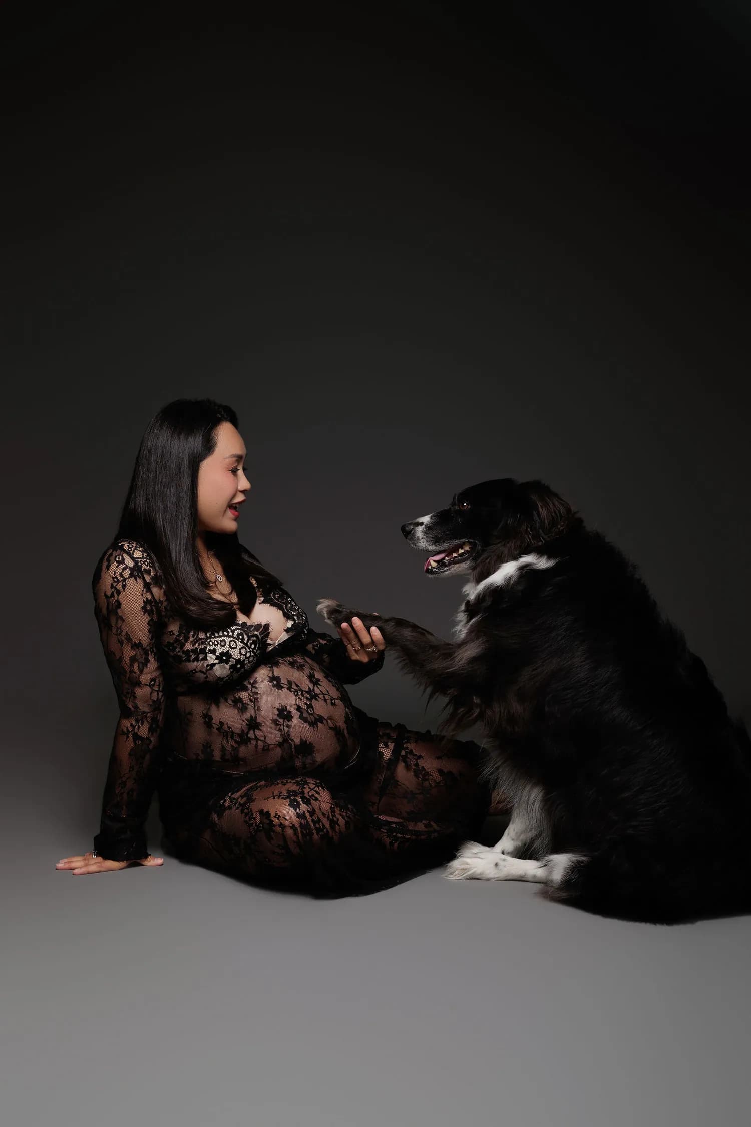 A pregnant woman in black lace sitting on the studio floor, holding the paw of a black and white dog