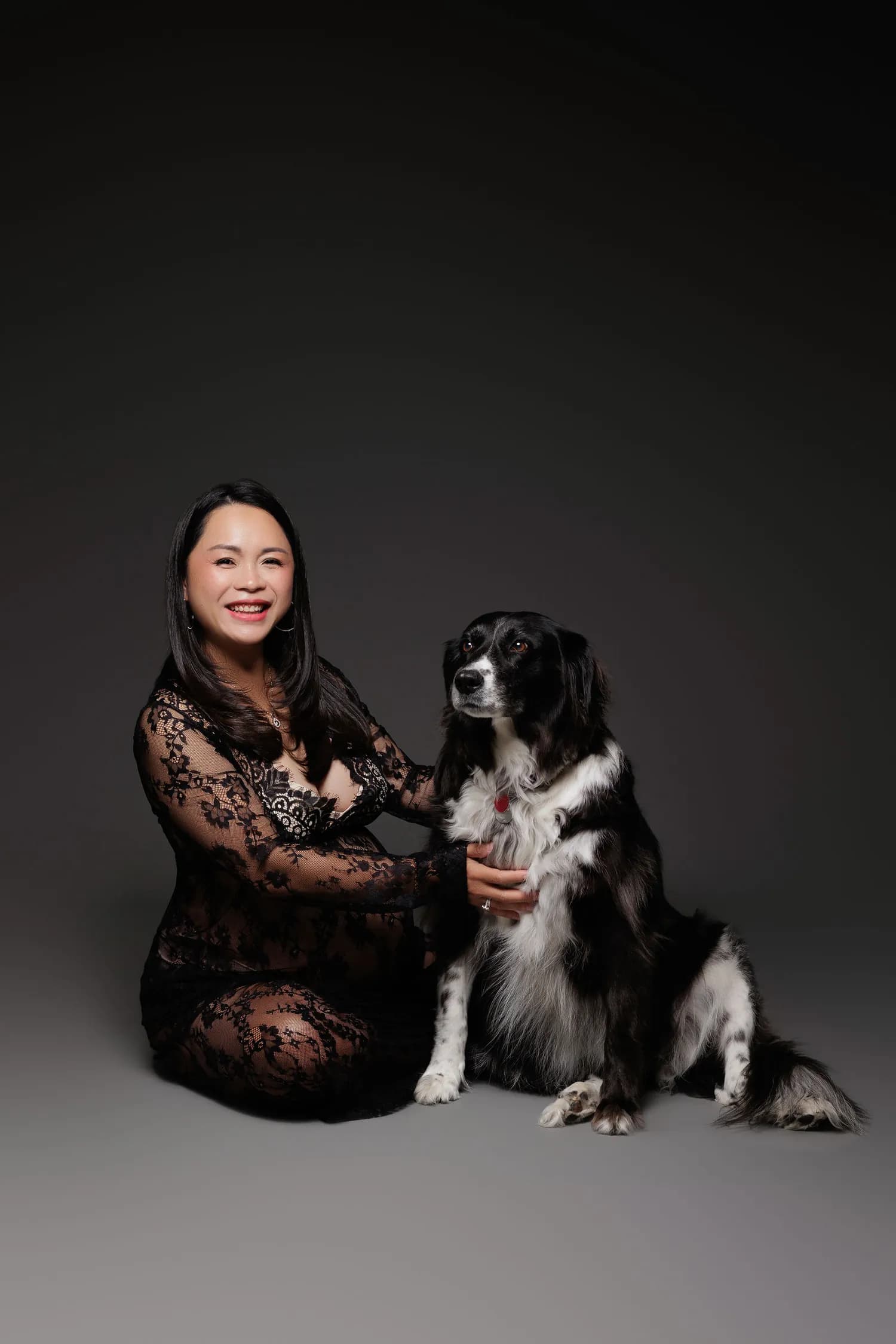 A smiling pregnant woman in black lace kneeling beside a black and white dog