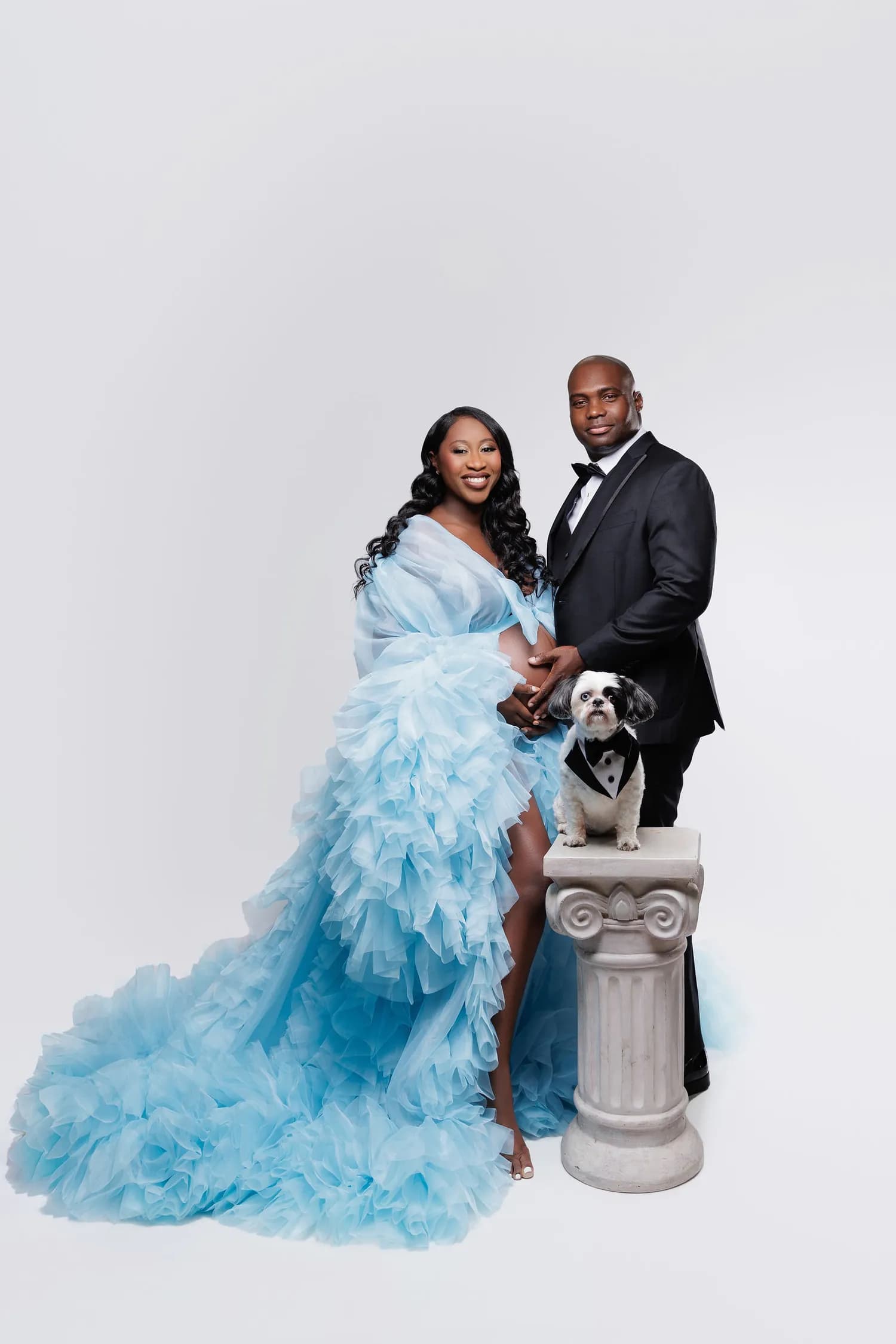 An expecting couple in formal wear with a small dog in a tuxedo on a white pedestal