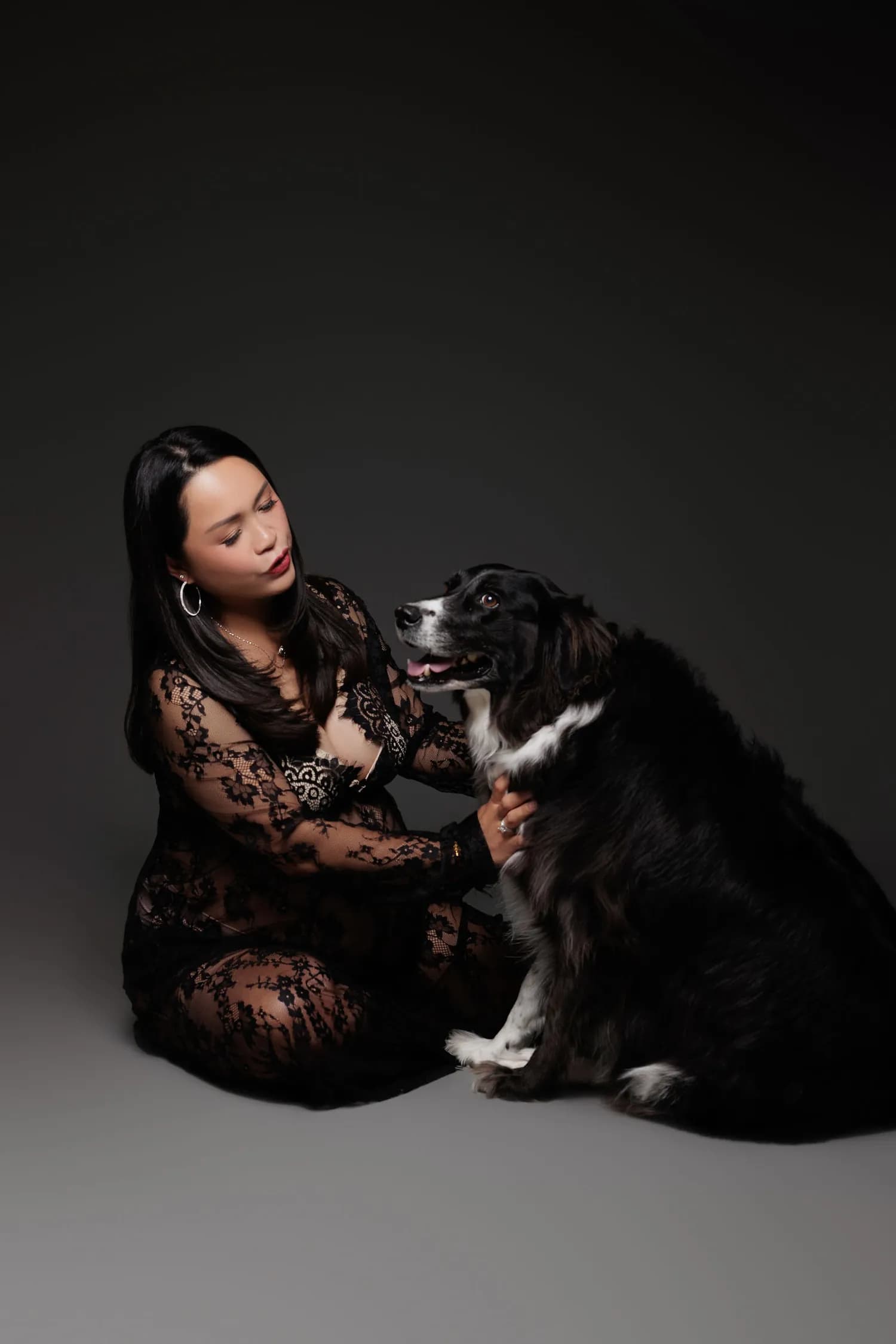 A pregnant woman in black lace holding a large black dog against a dark backdrop