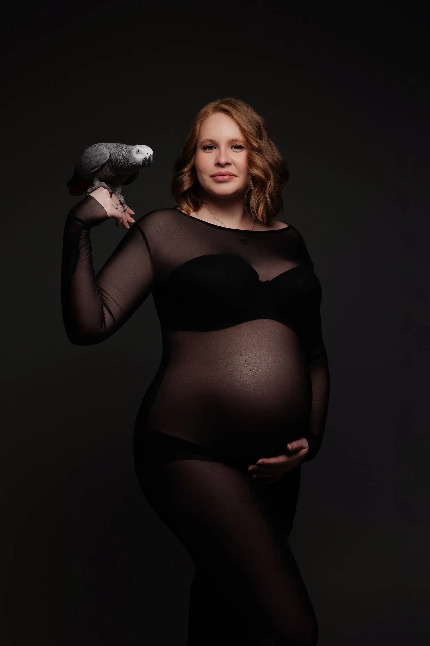 A pregnant woman in a sheer black gown with an African gray parrot perched on her hand