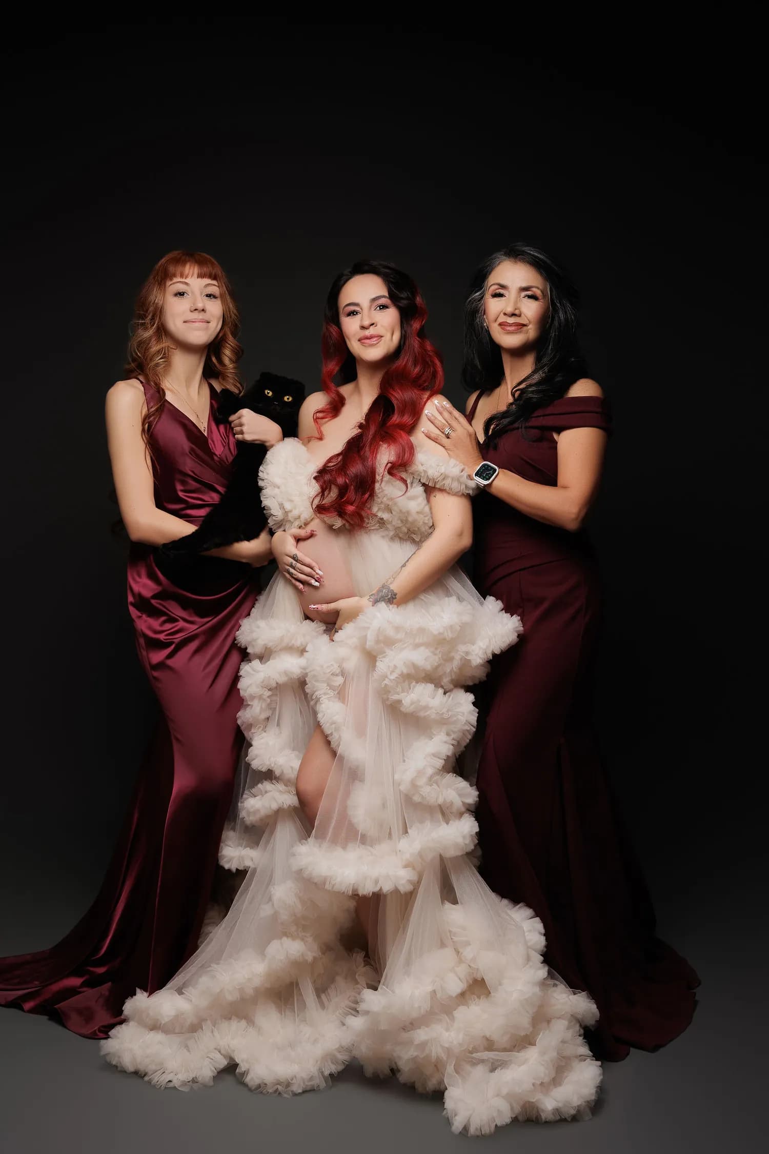 A pregnant woman in a blush ruffled gown holding a black cat, with two women in burgundy beside her