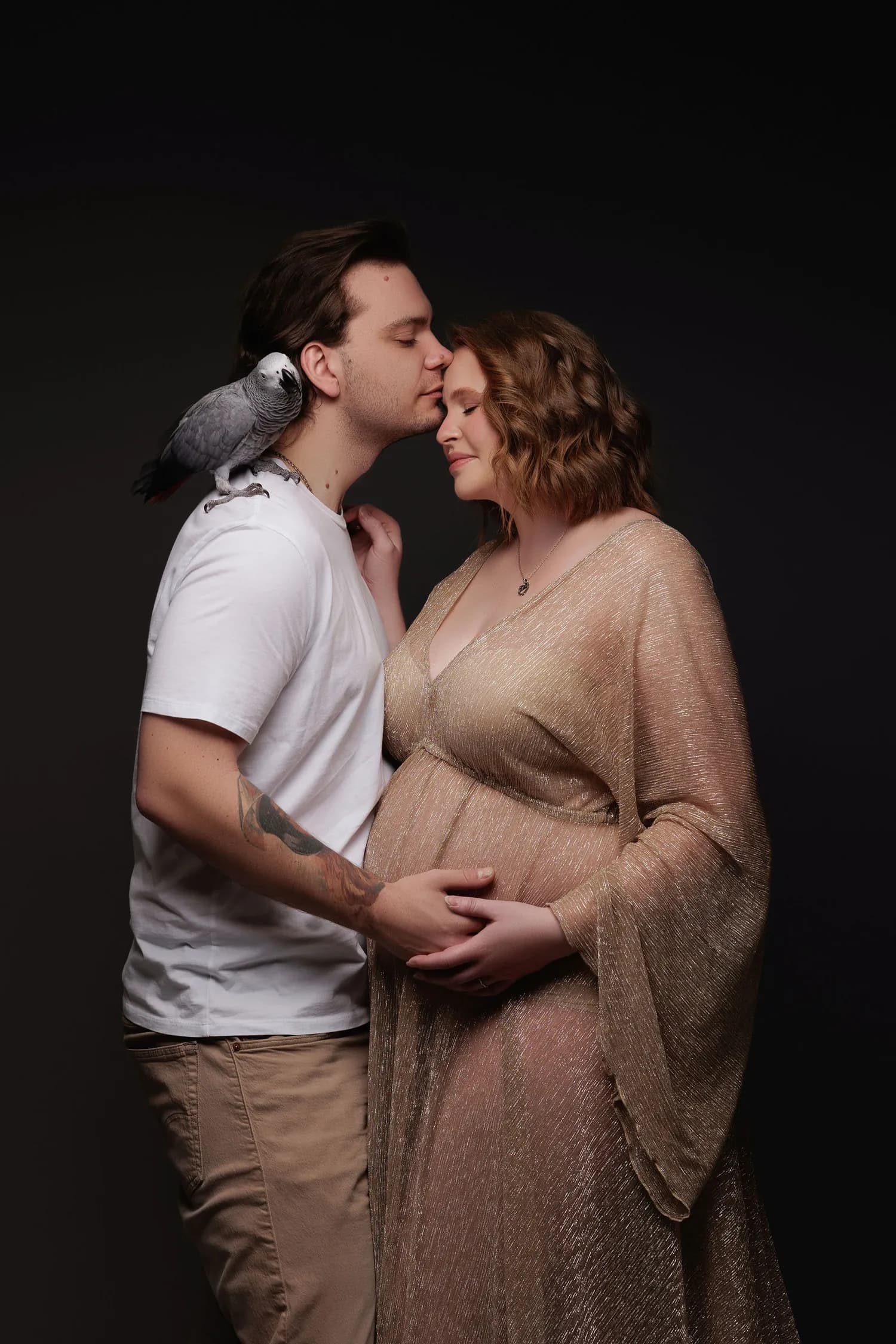 A man kissing a pregnant woman's forehead, an African gray parrot on his shoulder