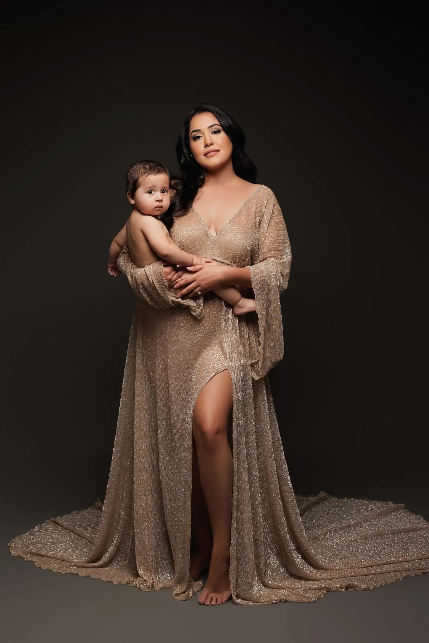 A mother in a shimmering champagne gown with a long train holds her baby at her side against a dark backdrop