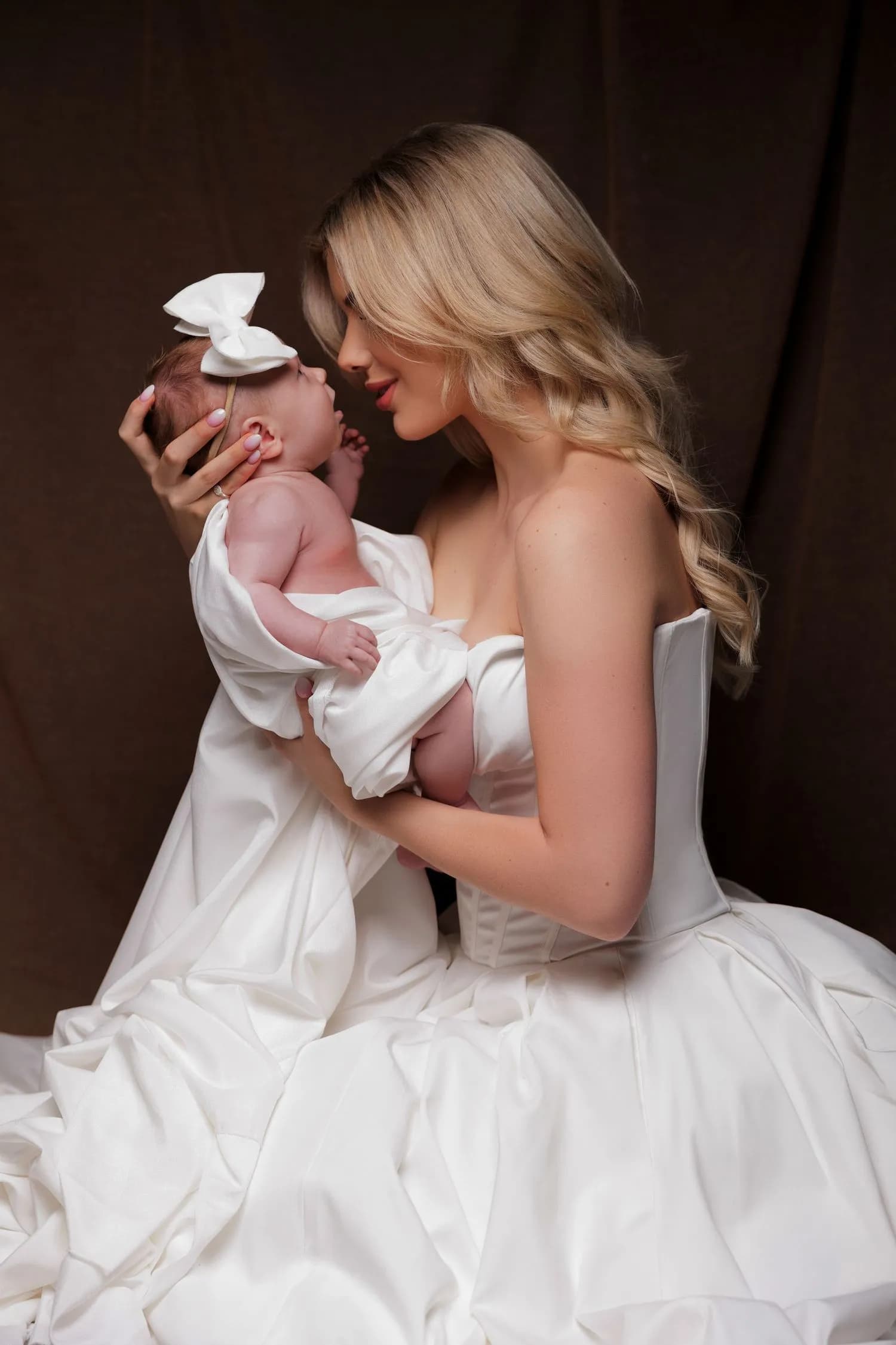 A mother in a white strapless gown lifts her baby toward her face; the baby wears a large white bow and is loosely wrapped in white fabric