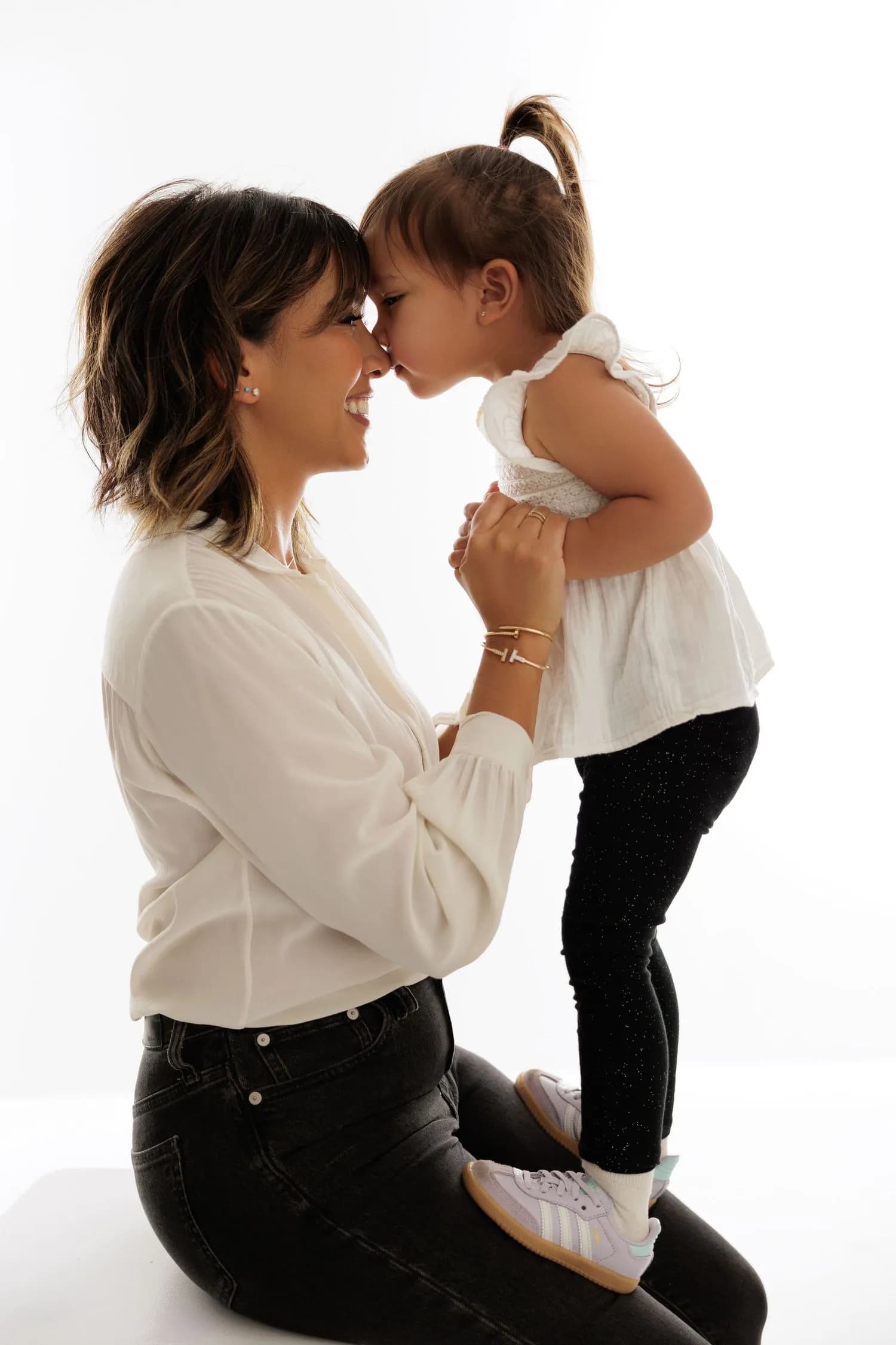A mother in a white blouse kneels and holds the hands of her young daughter, who stands on her lap and touches noses with her, against a white background