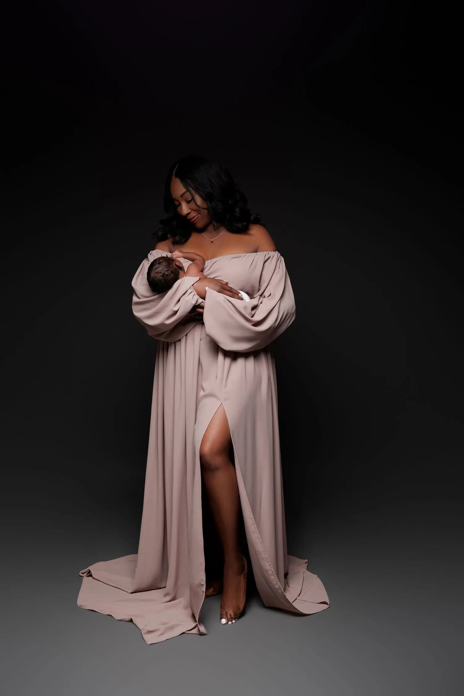 A mother in a long off-the-shoulder taupe gown with full sleeves stands barefoot against a dark backdrop, holding her baby close, the baby's face turned in toward her chest