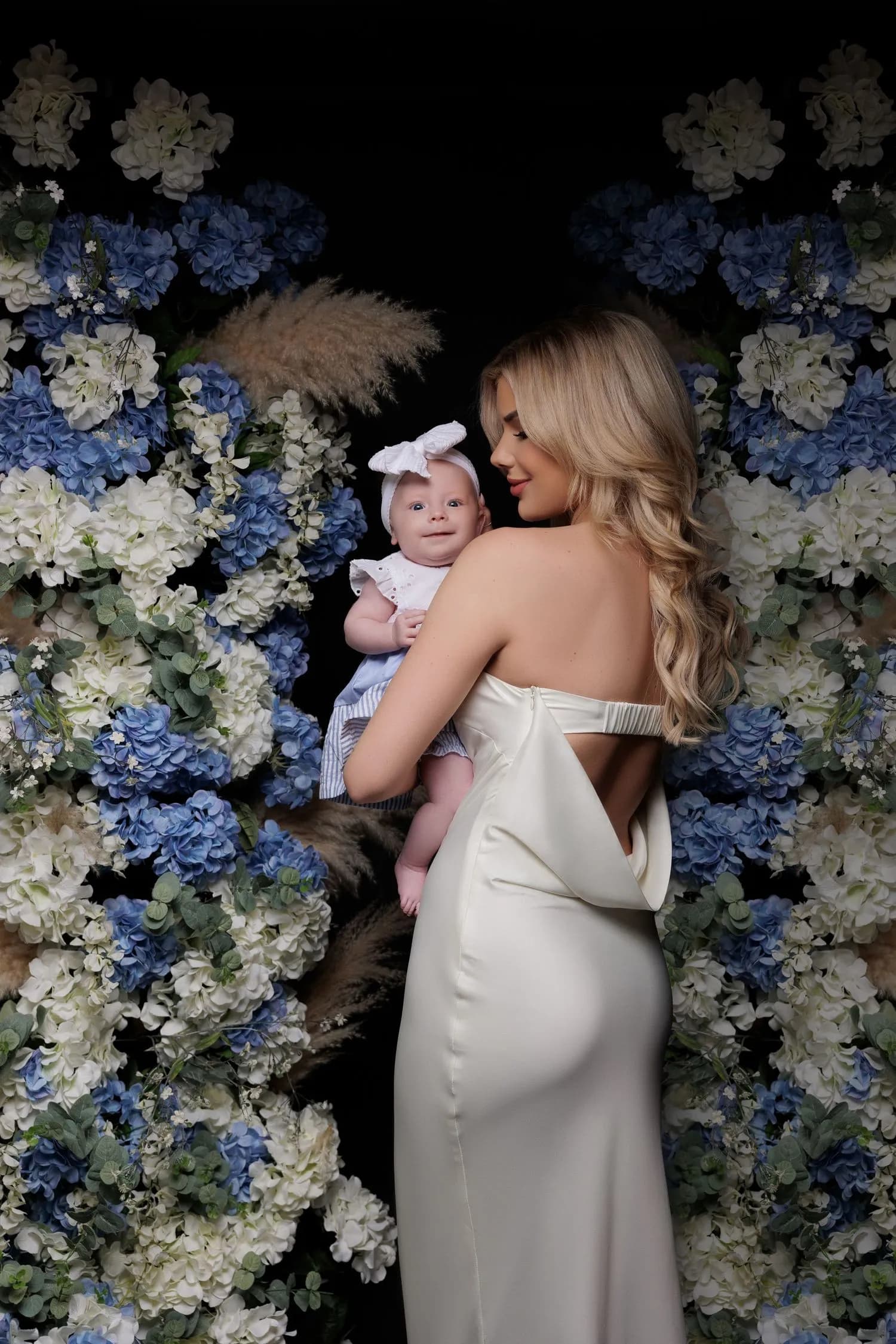 A mother in an ivory satin gown holds her smiling baby at her shoulder in front of a wall of blue and white hydrangeas