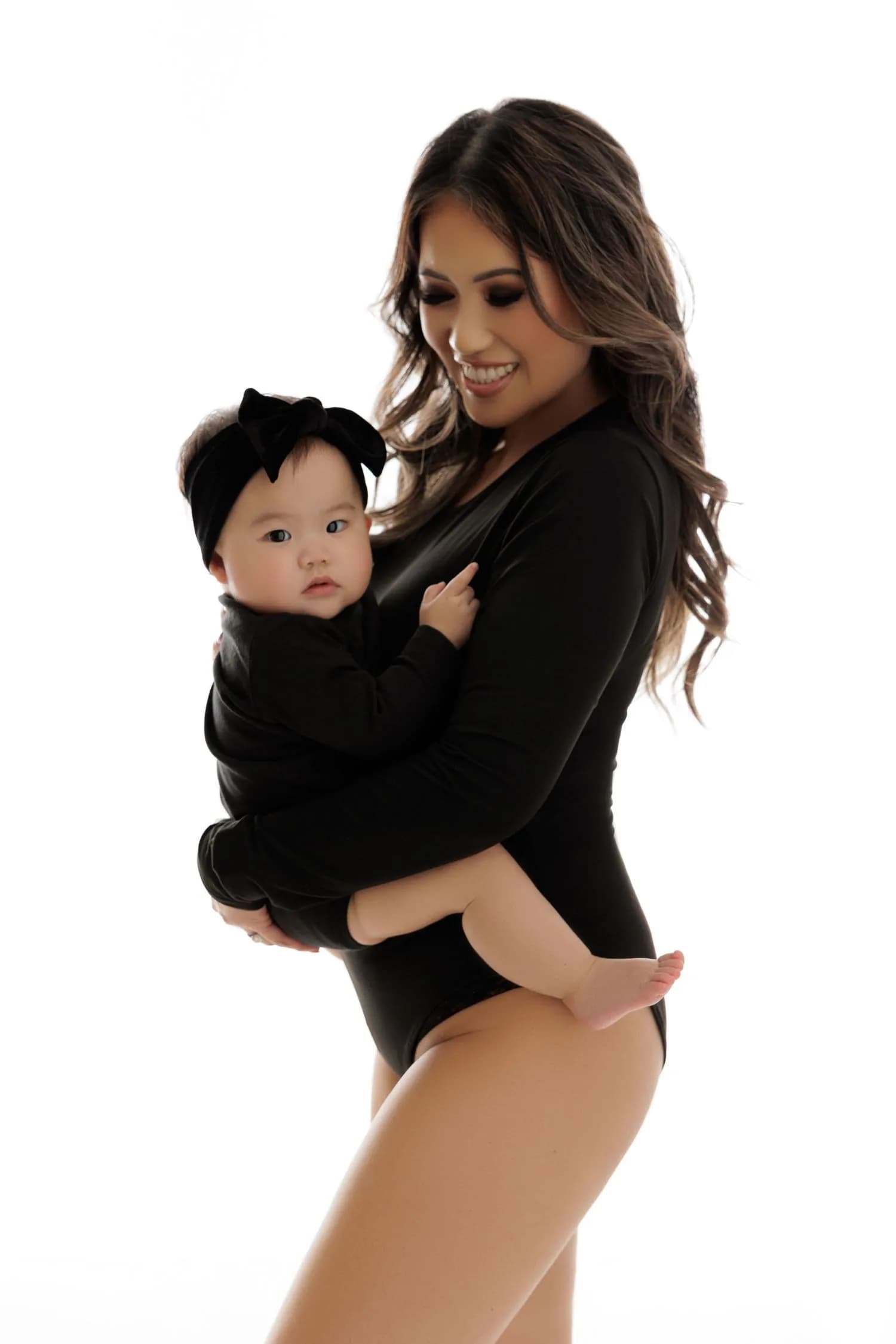 A smiling mother in a black bodysuit holds her baby, dressed in black with a black bow headband, against a white background