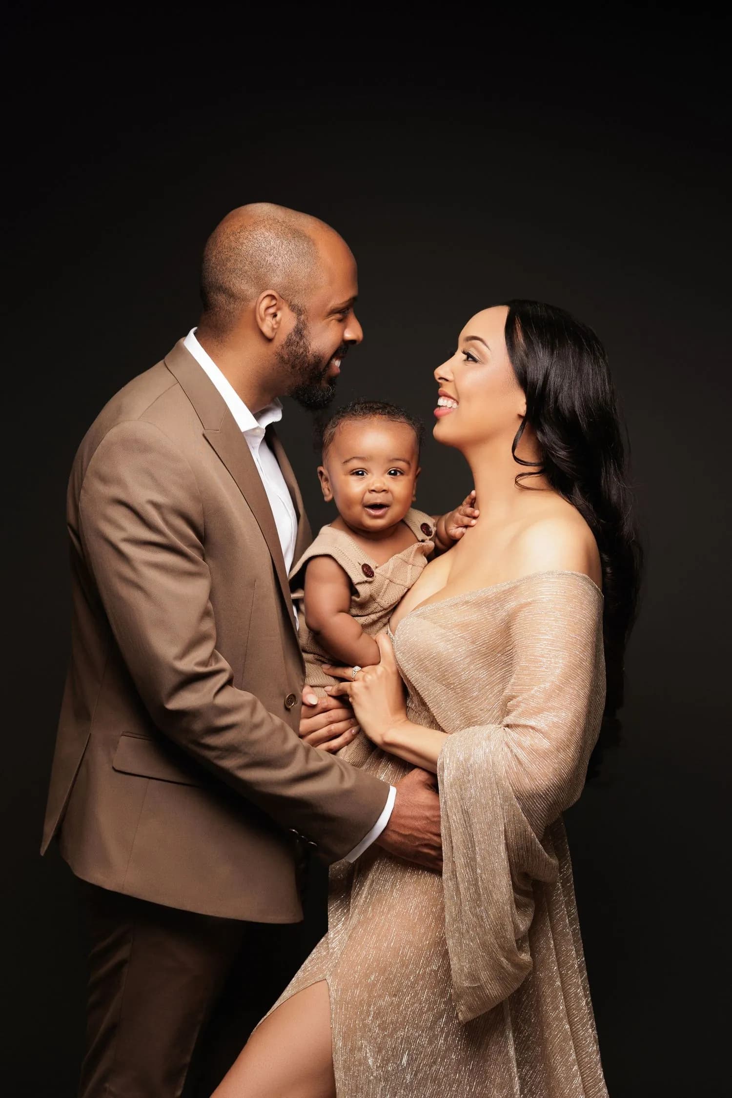 A father in a tan suit and a mother in a shimmering off-the-shoulder gown smile at each other while she holds their smiling baby between them