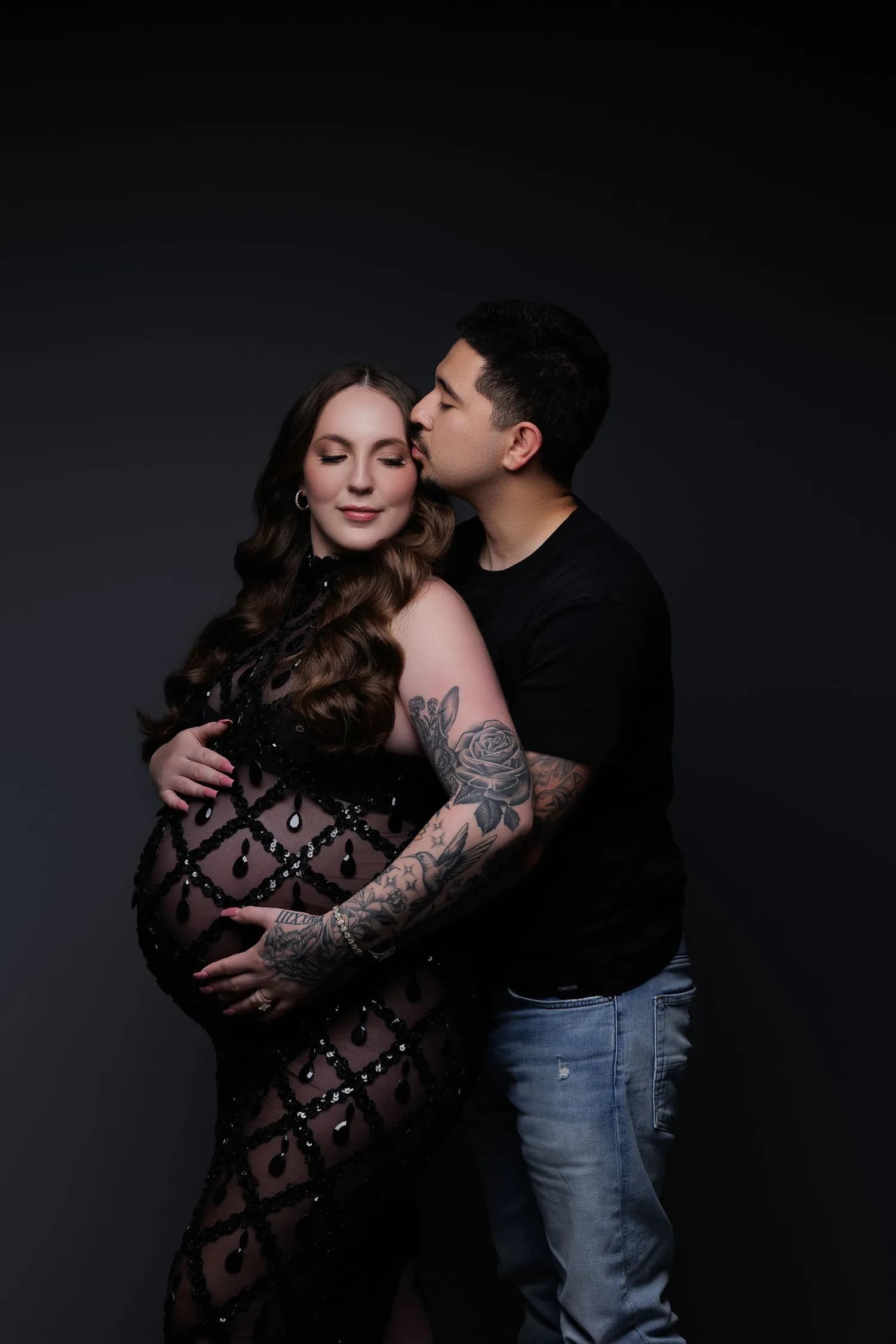 Partner holding a pregnant woman in a black lace gown and kissing her cheek